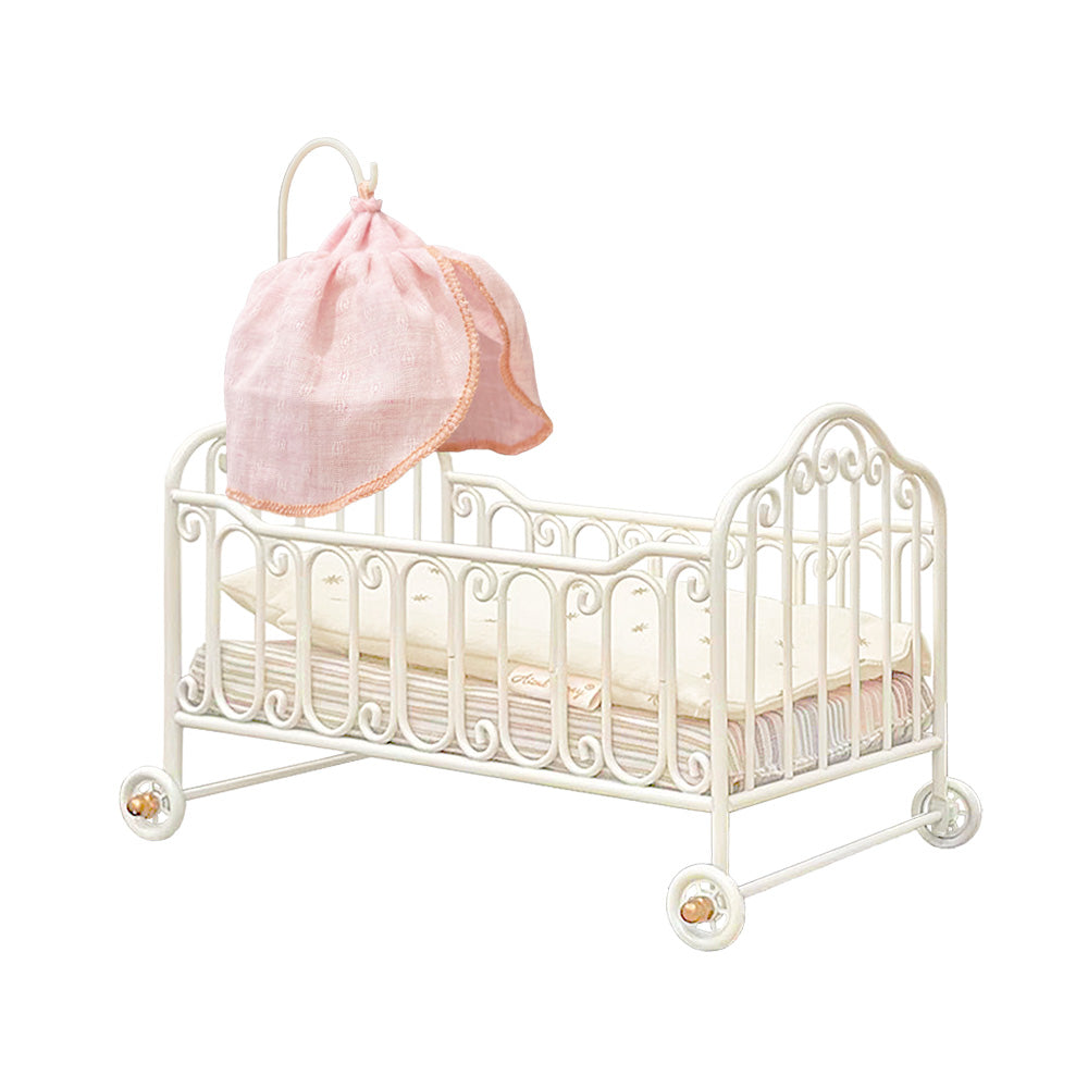 Cream-white metal miniature baby cot with hand-bent scrollwork, rolling wheels, cotton bedding and a pink gauze canopy on a removable curved support, on a white background