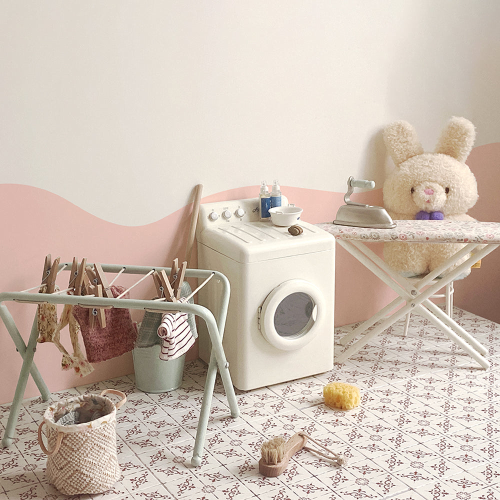 Off-white miniature washing machine in a doll laundry scene with drying rack, ironing board and iron, basket and toy bunny used as props